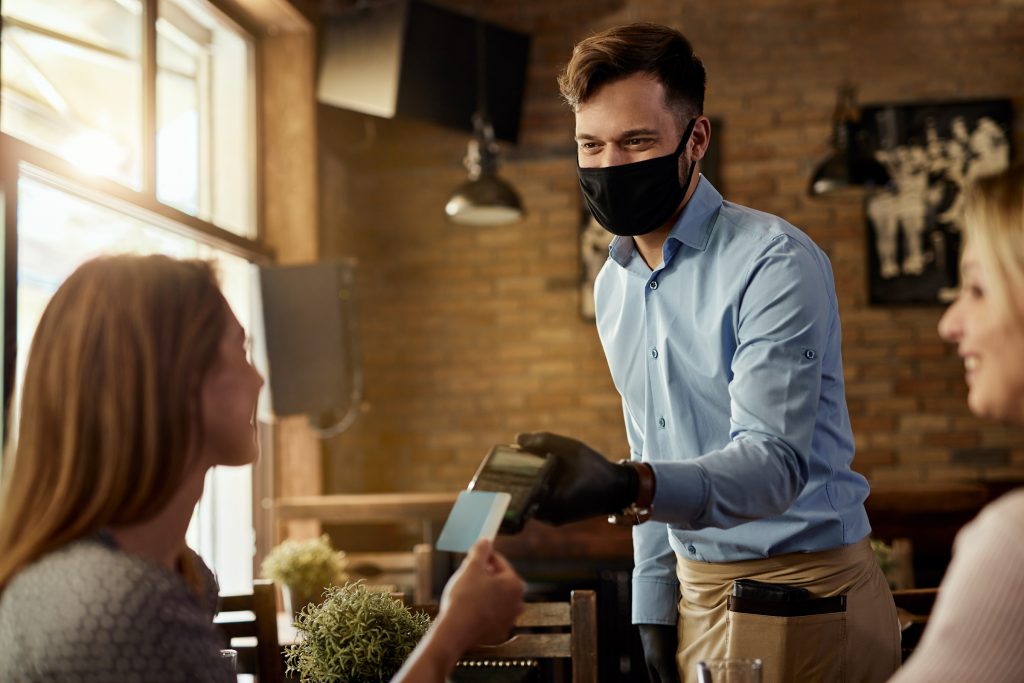 male customer making contactless payment to a waiter who is wearing protective face mask in a cafe.